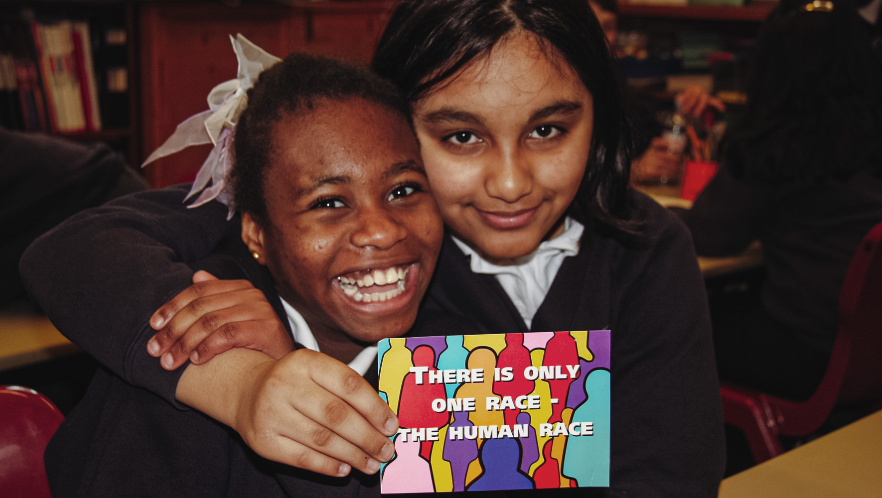 Two schoolgirls in a classroom are smiling and holding a postcard. It reads: 