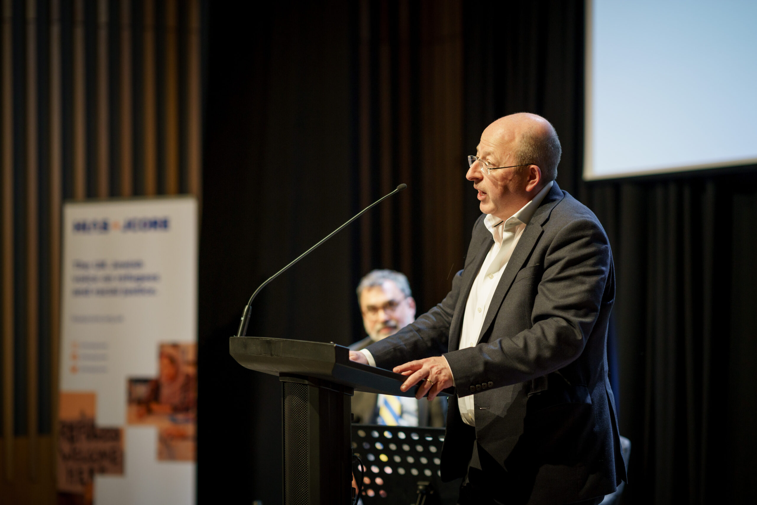 HIAS+JCORE's chair Adam Rose speaks at a lectern at the launch event. In the stage background stands Mark Hetfield, the HIAS President and CEO.