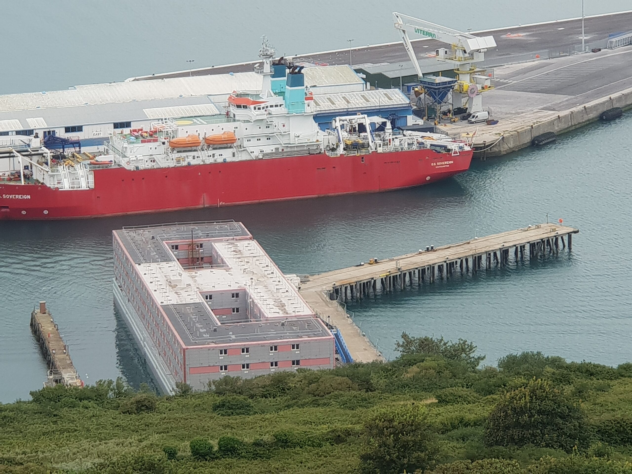 Image shows the docked Bibby Stockholm barge, taken from a hillside over looking the sea.