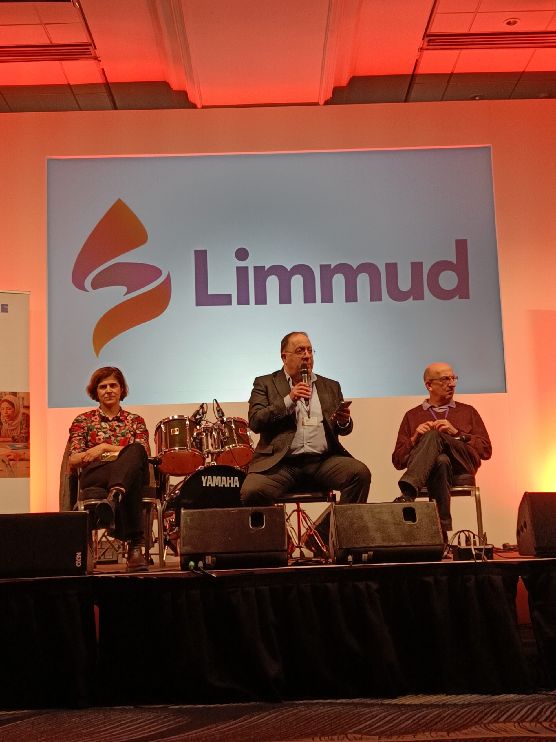 David Mason speaks on stage at Limmud. He is sat holding a microphone. To his left is Sarah Sackman, and right, Michael Wegier (Board of Deputies), both also sat.