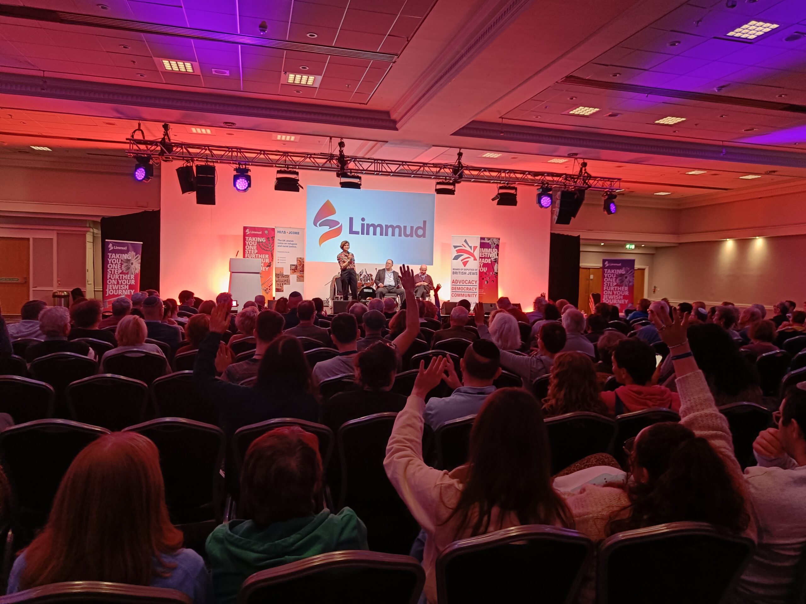 Image from the back of the main room at Limmud. Rows of people are sat in front of the stage, where Sarah Sackman is speaking. She is stood in front of a projection of 'Limmud' on a screen.