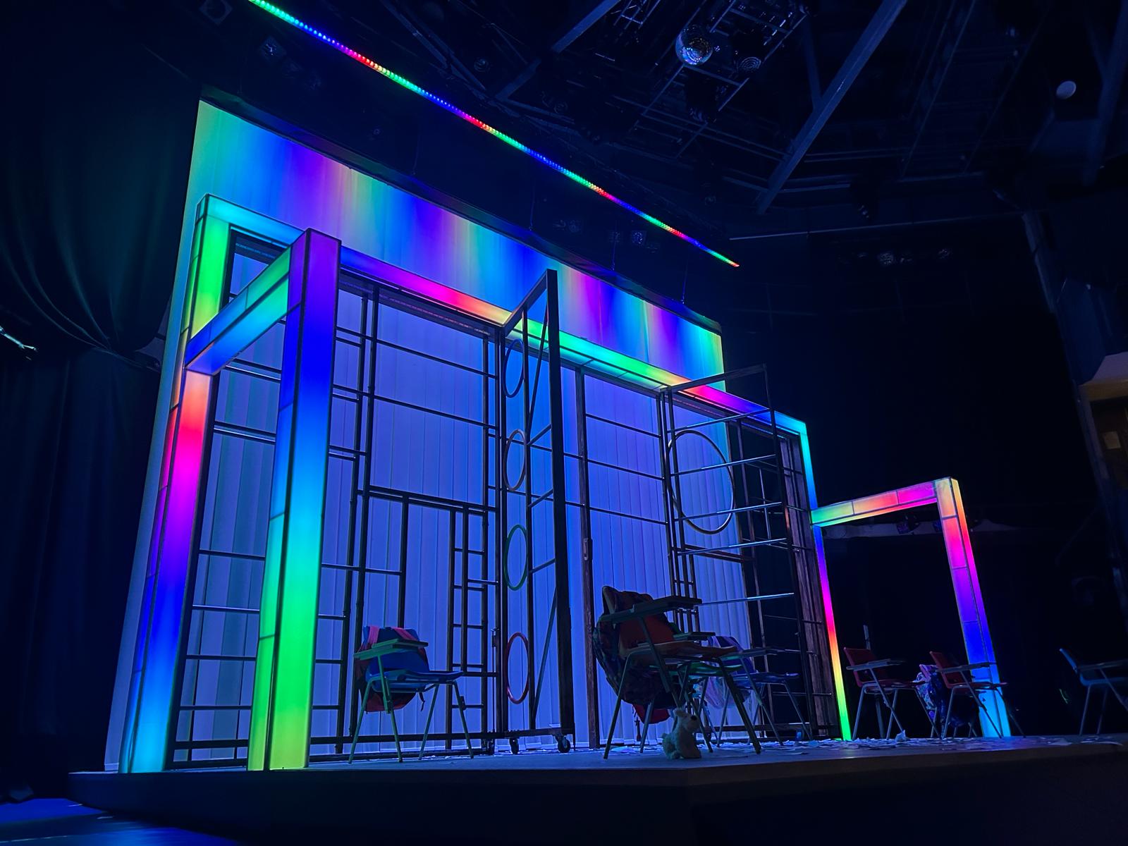 Stage at the Boy at the Back of the Class play. There are chairs across, and the stage is backlight with green, blue and orange lights.