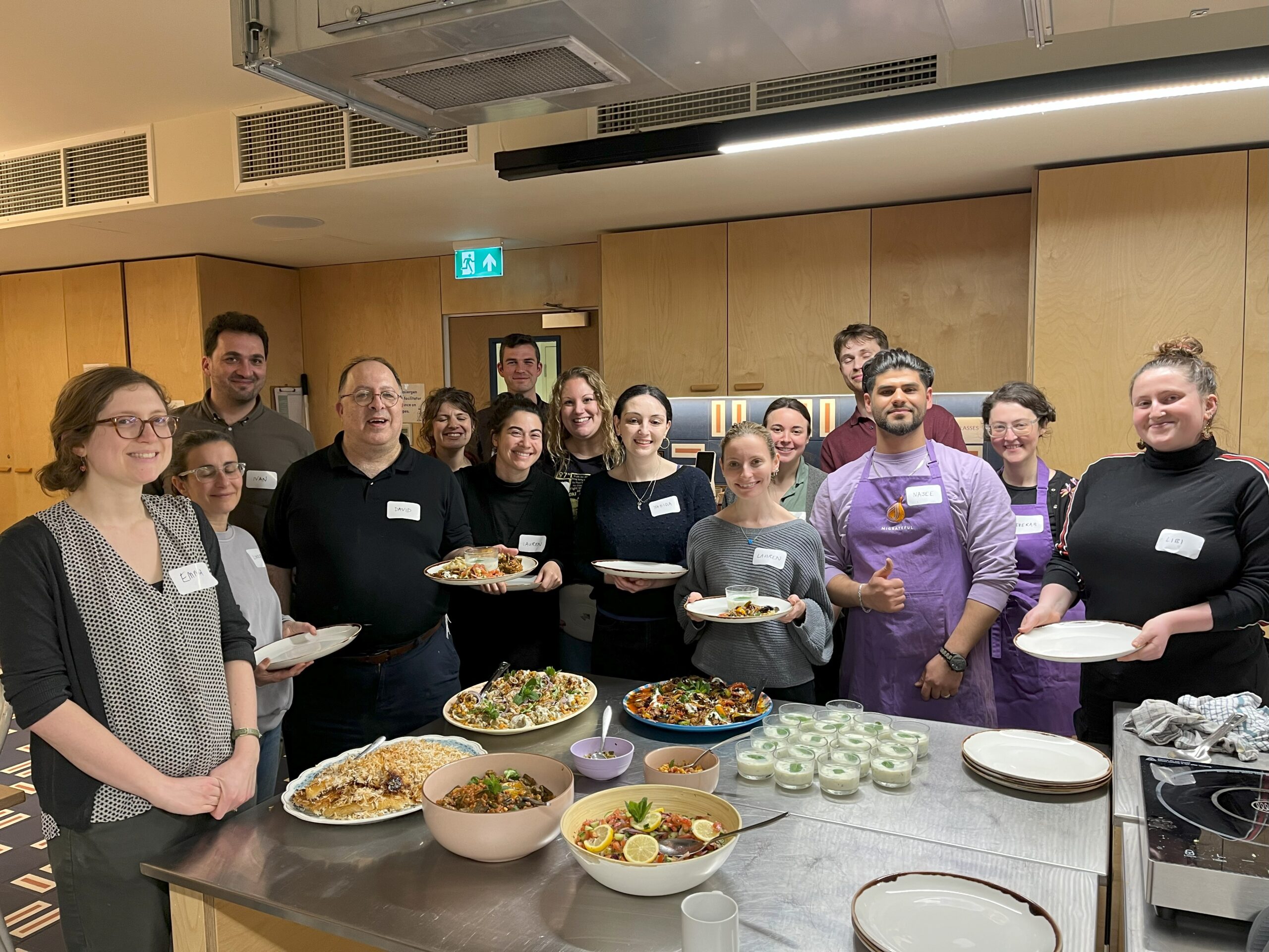 Image shows a group of people looking towards the camera. They are stood in a kitchen in front of a set of plates of food.